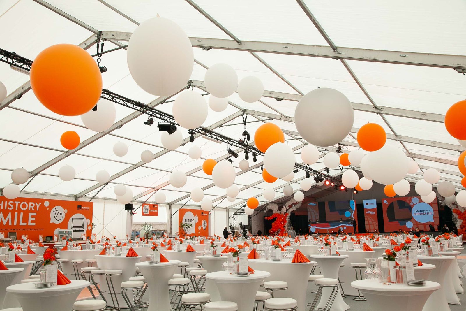 Firmenjubiläum im Zelt mit orangen und weißen Ballonen, Stehtischen und Barhockern